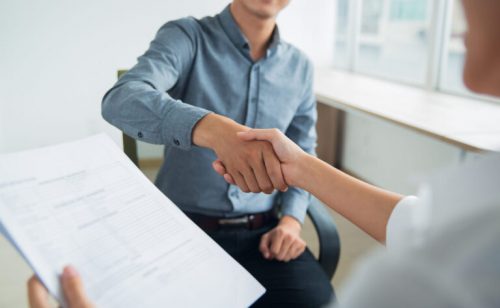 Smiling Asian businessman wearing shirt sitting in office and shaking hand of female partner. Woman holding document