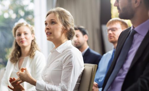 Business people sitting and watching presentation in conference room. Young businesswoman telling something to speaker and gesturing. Speaker is out of view.